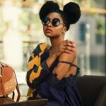 Fashionable woman in a chic outfit and sunglasses sitting indoors with a stylish handbag.